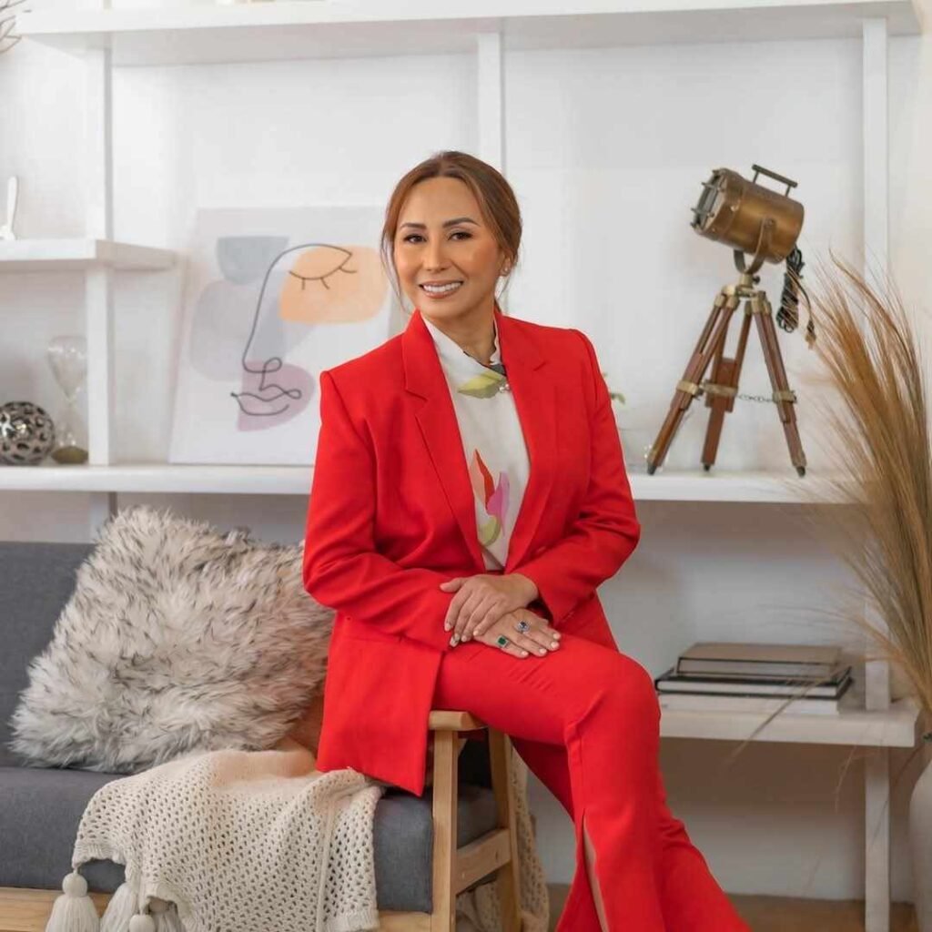 A woman in a bright red suit sits on a gray couch in a stylish room with white shelves, books, decorative items, and a vintage tripod camera in the background.